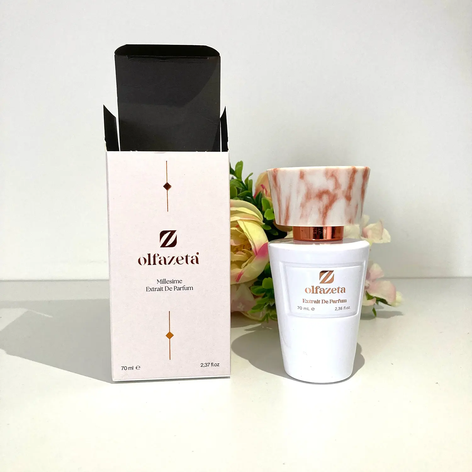Flacon de parfum Olfazeta Millésime Extrait de Parfum 70 ml avec bouchon effet marbre rose, présenté à côté de son étui élégant blanc et doré, sur fond clair avec décoration florale