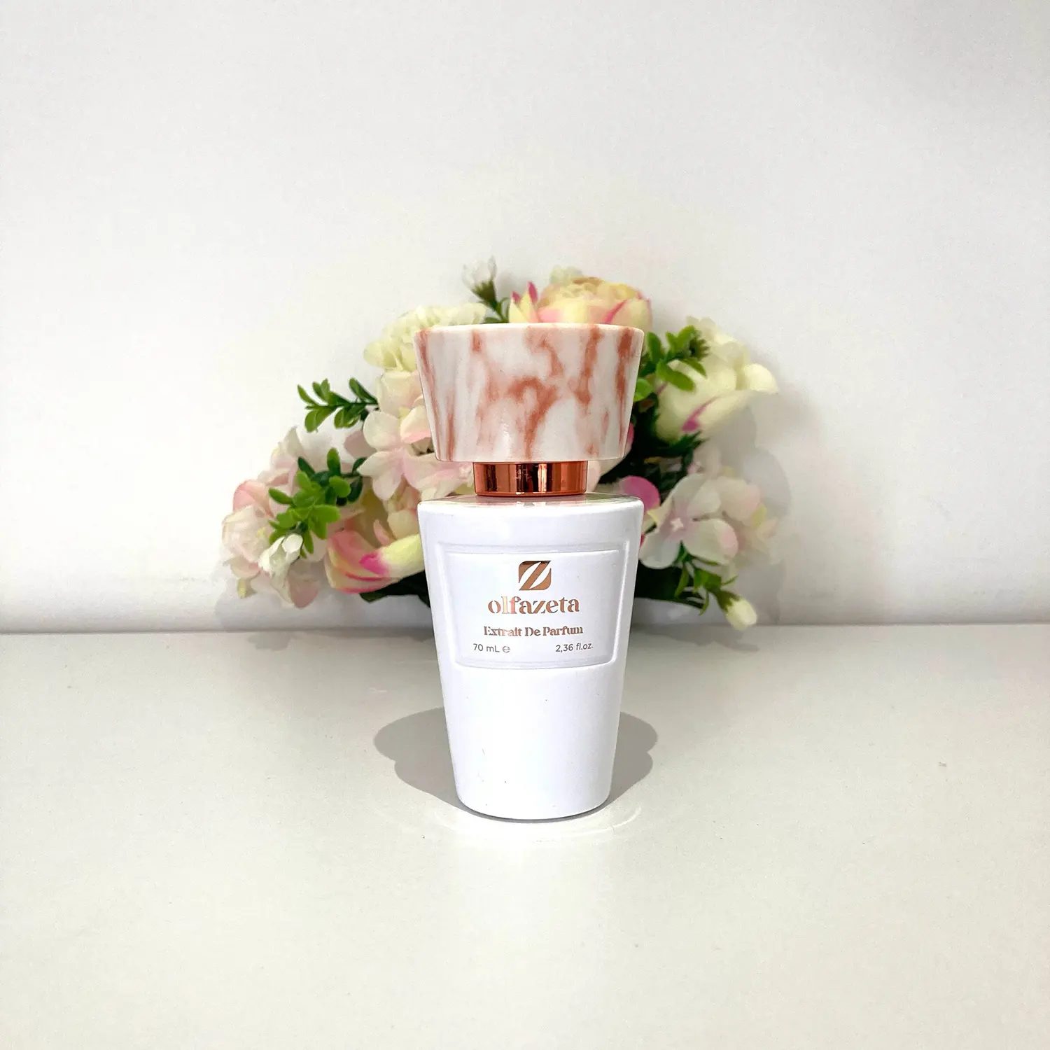 Flacon de parfum Olfazeta Extrait de Parfum 70 ml posé devant un fond floral, avec bouchon marbré rose et doré, présenté en gros plan pour en valoriser le design élégant
